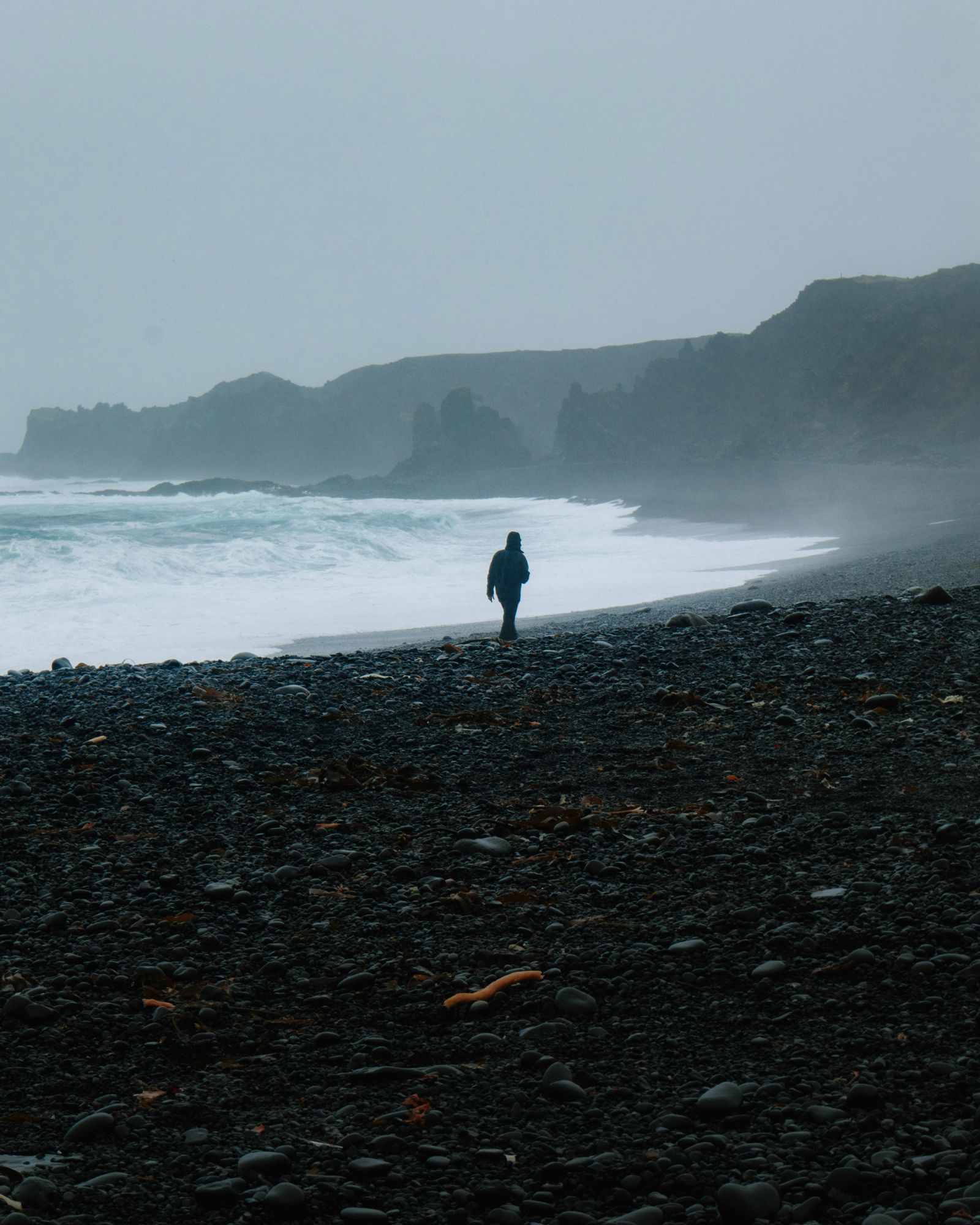 Pessoa caminhando sozinha por uma praia de pedras escuras, diante do mar agitado e de falésias na neblina